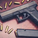 Scadenza del porto d’armi e stato d’emergenza: che succede? Scadenza del porto d’armi e stato d'emergenza: pistola Glock su tavolo di legno con munizioni